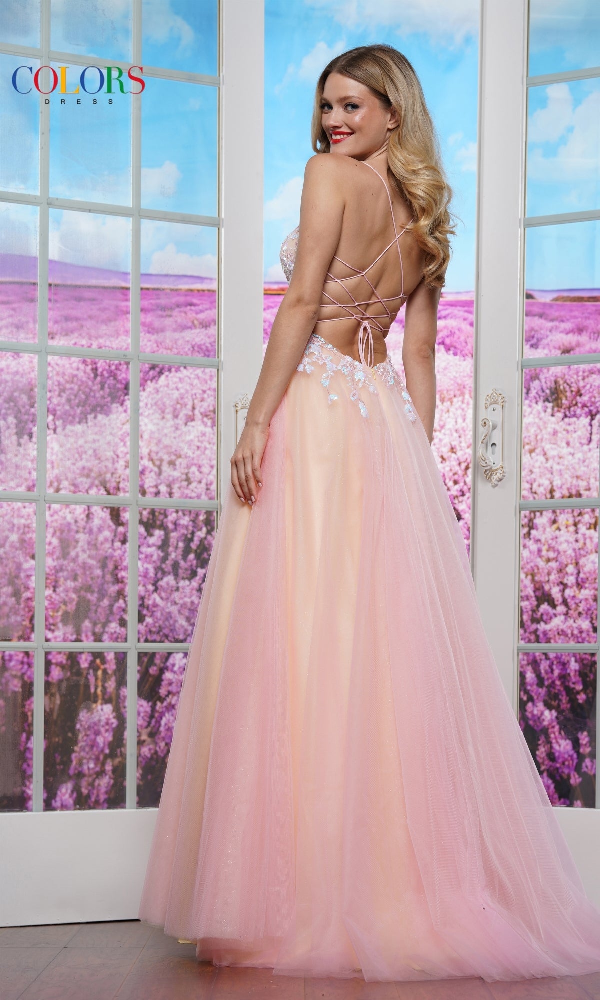 Wearing the Strappy-Back Prom Ball Gown: Colors Dress 3497, a woman stands before glass doors with a field of purple flowers outside, her long A-line dress featuring a sheer bodice and floral details.